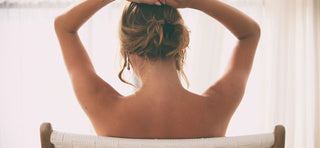 Nude woman sitting on chair facing away from camera with arms touching blonde hair